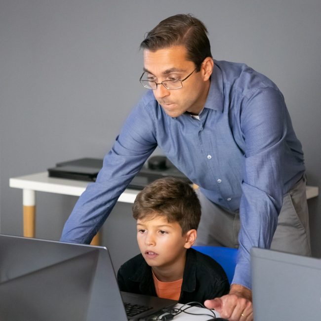 Content teacher checking task and standing behind pupil Content teacher checking task and standing behind pupil. Focused boy sitting at table in school, waiting for assessment from tutor and looking at screen. Knowledge and digital education concept